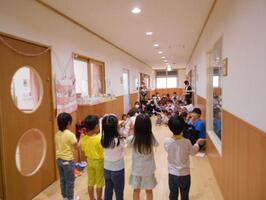 部屋の前の廊下で0歳児～歳児が集まり、その前で5歳児が手遊びを披露しています