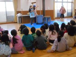 保育教諭が餅つきの道具である杵を持ちながらそれについて説明し、園児が並んで座って聞いています。