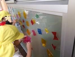 2歳児の子どもたちが、窓にいろいろな色のセロハンを貼って遊んでいます。