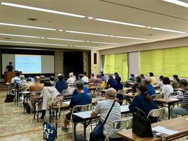 会場前方にスクリーンがあり、講義資料が投影されている。講師はスクリーンの左にいる。受講者用に机と椅子があり、講師の話を聞いている。会場の左斜め後ろから撮影している。