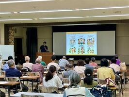 会場前方にスクリーンがあり、講義資料が投影されている。講師はスクリーンの左にいる。会場の右斜め後ろから撮影した写真。