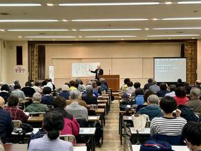 舞台上の中央で講師がマイクを持って話している。その前に参加者用の机と椅子があり、多数の参加者が講師の話を聞いている。