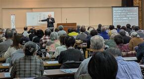 舞台上の中央で講師がマイクを持って話している。その前に参加者用の机と椅子があり、多数の参加者が講師の話を聞いている。写真の左上には、講演内容の要約文を投影したスクリーンがある。