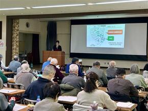 中央に資料を投影したスクリーン。左の演台にいる講師がマイクを持ち、中村地域について話している様子。