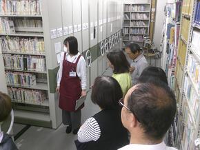 画像左側に周密書架があり、職員がその前に立って書庫にある資料の説明をしている。
