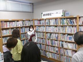 画像全体に教育情報コーナーの本棚があり、職員が中央に立って説明している。