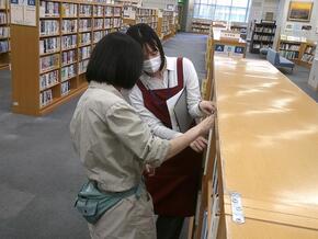 画像右側に本棚がある。その前に職員と受講者が一人ずつ立ち、書架整理をしている。